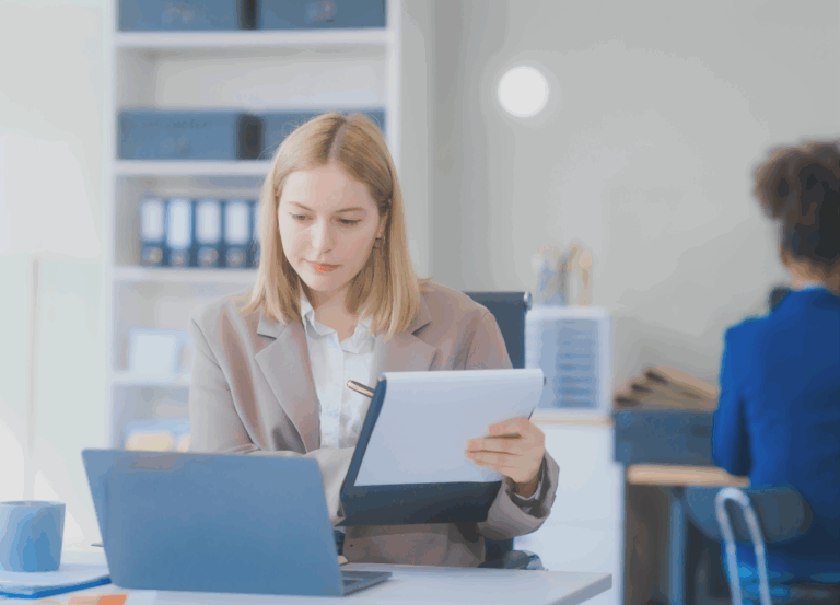 Woman holding a notebook and looking at a laptop.
