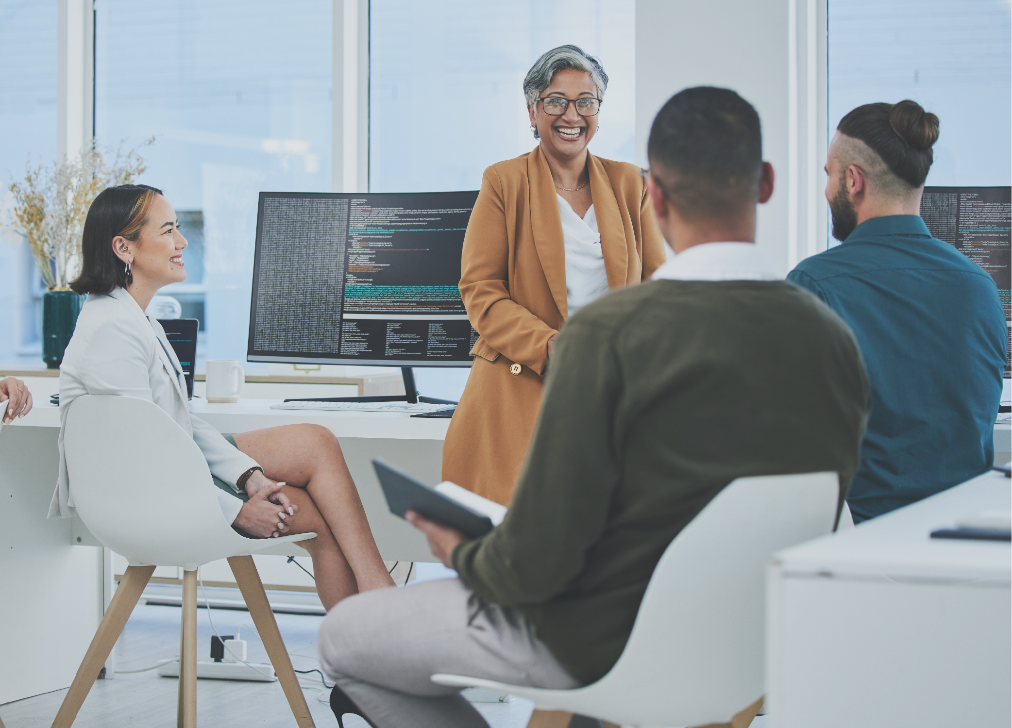 Four employees having a meeting with a computer monitor full of data behind them.