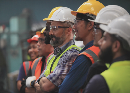 Seven employees all wearing safety vests and hard hats while looking at something.