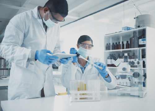 Two lab employees collaborating and working while wearing goggles and a mask.