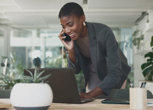 Woman on a phone while leaning over a laptop and smiling.